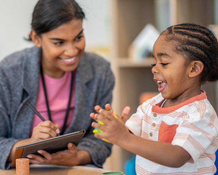 Smiling therapist taking notes on a tablet while a young child laughs and gestures during a speech therapy session.