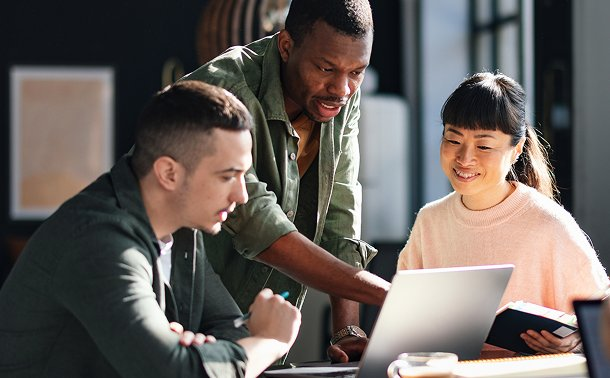 Three diverse coworkers reviewing content on a laptop together at a sunlit office table during a team discussion.