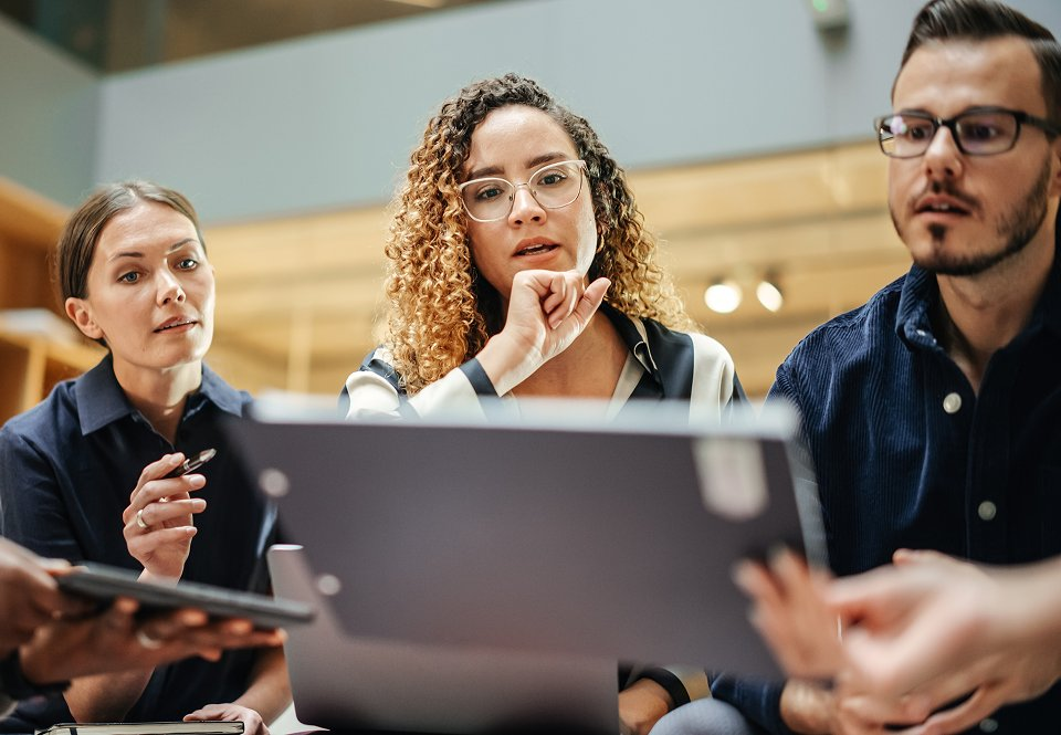 Three diverse coworkers gathered around a laptop in focused discussion, with one woman thinking thoughtfully in the center.