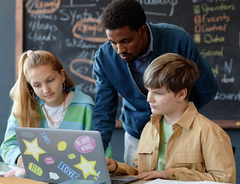 Male teacher leaning over to help two students working on a sticker-covered laptop in a classroom with chalkboard notes behind.