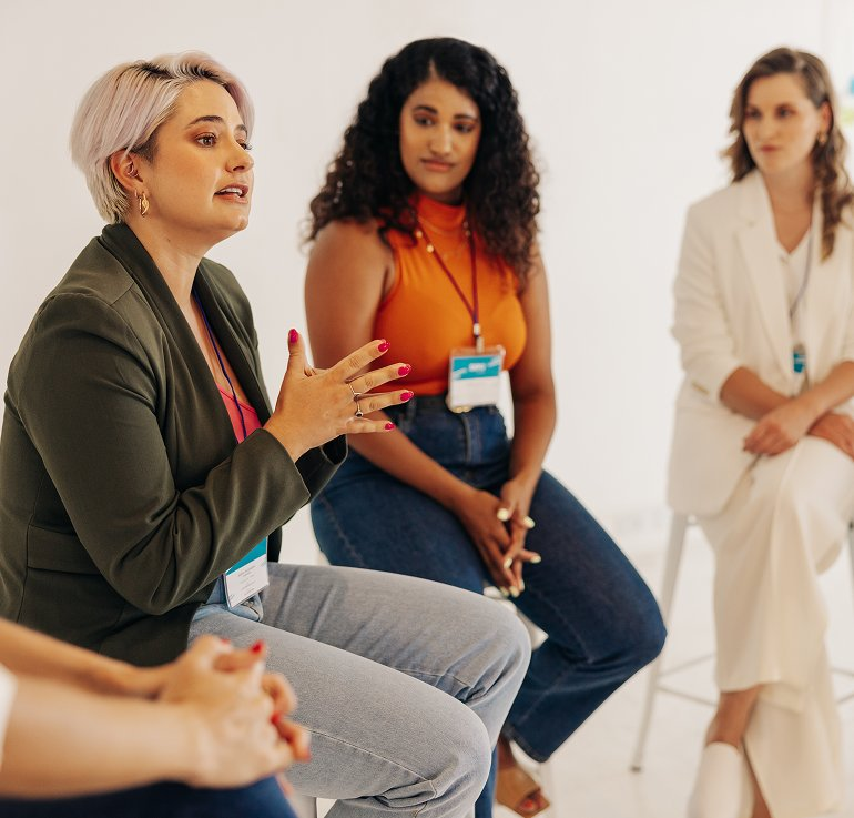 Diverse women seated in a circle wearing lanyards, with a woman in a green blazer speaking and gesturing during a group discussion.